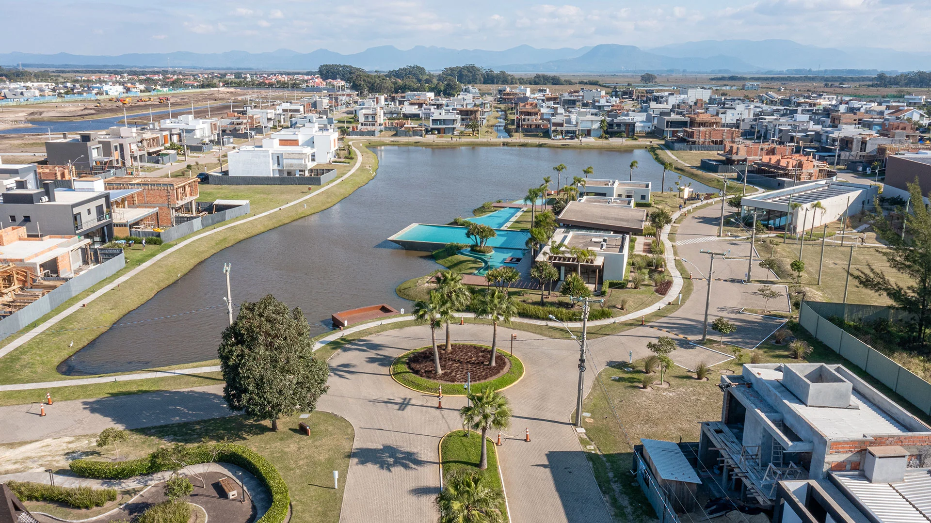 Foto aérea panorâmica do Blue Xangri-lá com a rotatória de palmeiras na entrada, o lago central, o clube ao fundo e a serra no horizonte