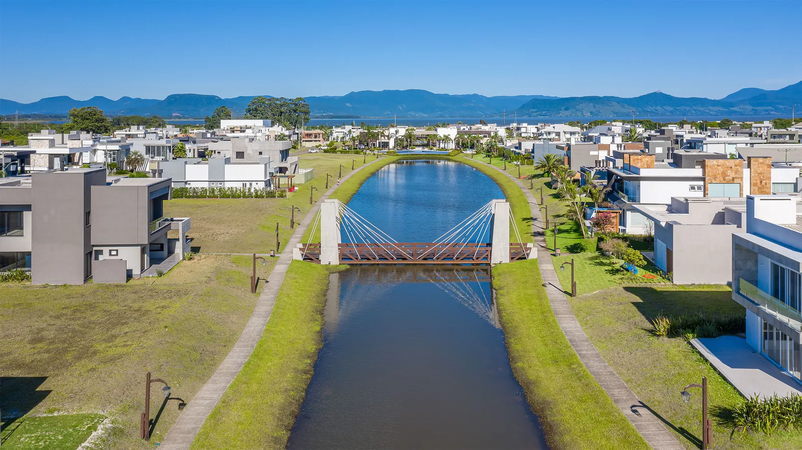 Ponte suspensa estaiada de pedestres sobre um canal interno do Capão Ilhas Resort, com casas dos dois lados e a lagoa e a serra ao fundo