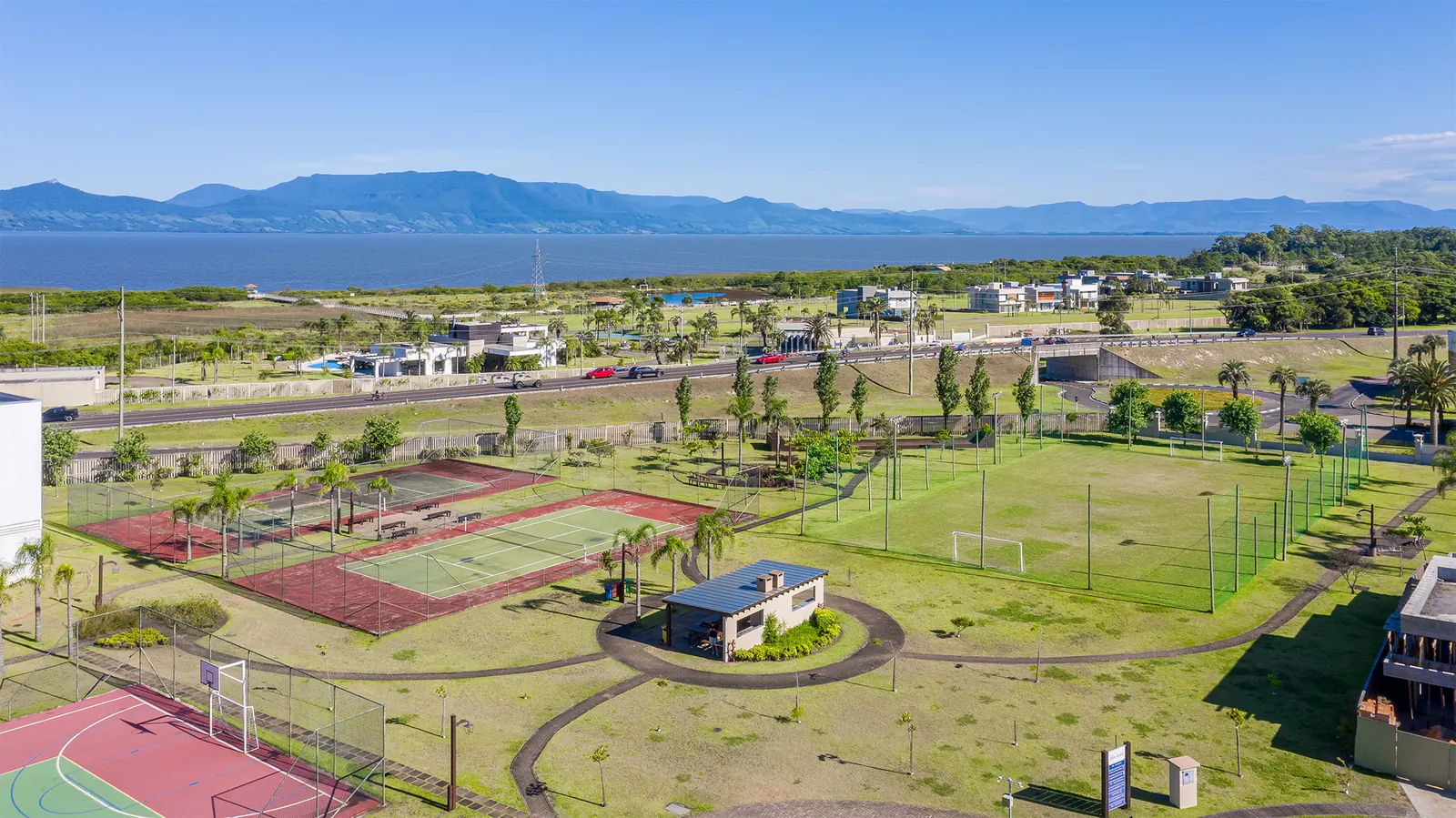 Foto aérea das quadras de tênis, poliesportiva e do campo de futebol society do Capão Ilhas Resort, com o ginásio coberto, a lagoa, a serra e aerogeradores no horizonte