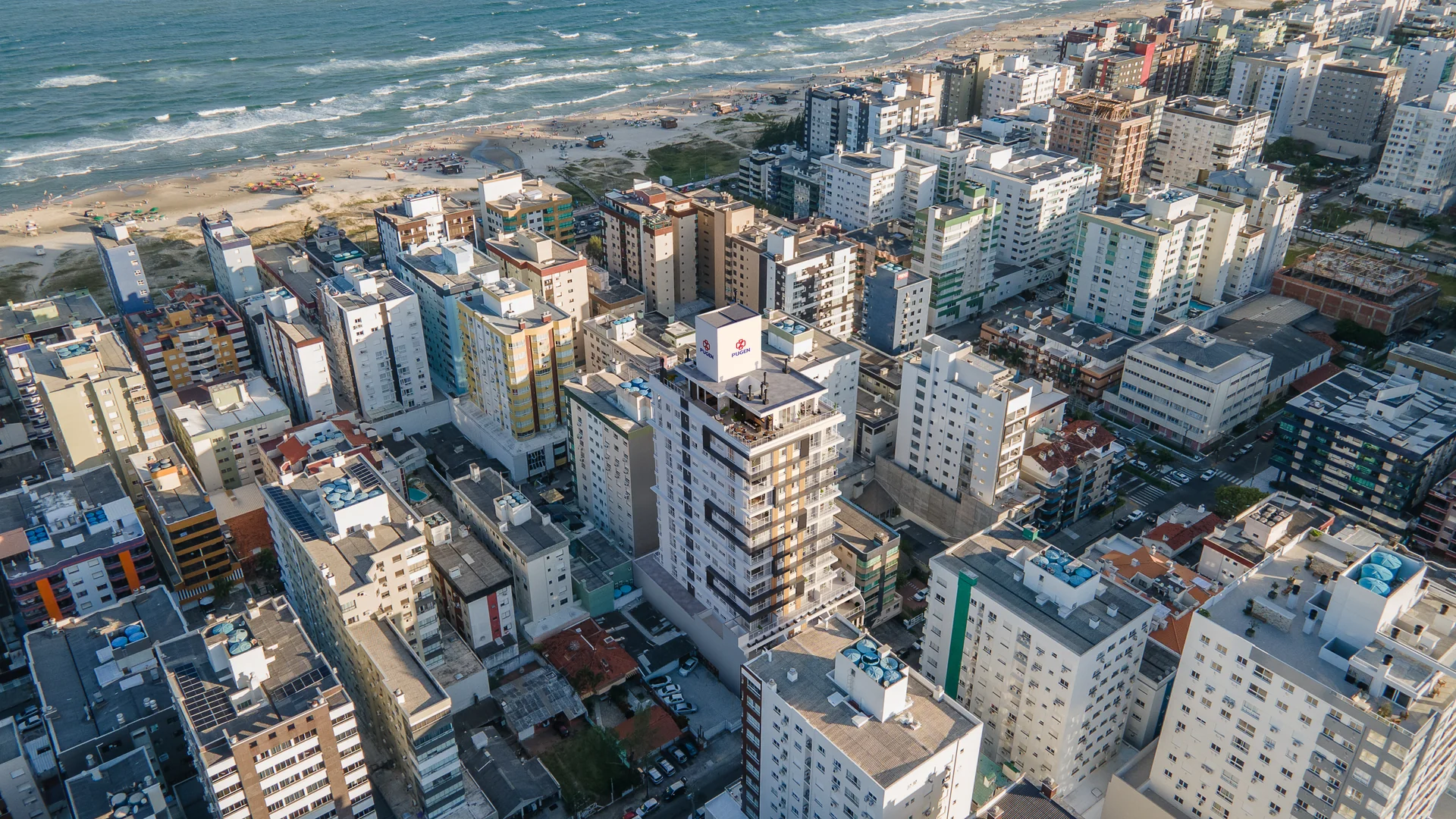 Perspectiva aérea do Centro de Capão da Canoa com a torre do Chapter28 Residence implantada na malha urbana e o mar ao fundo, a poucas quadras