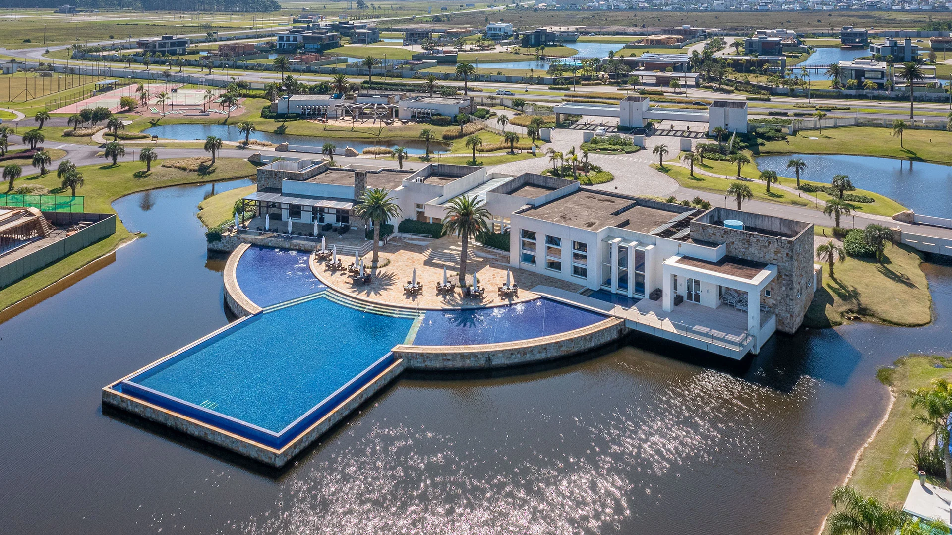 Foto aérea do club house do Enseada Lagos com a piscina de borda infinita avançando sobre o lago e deck com guarda-sóis