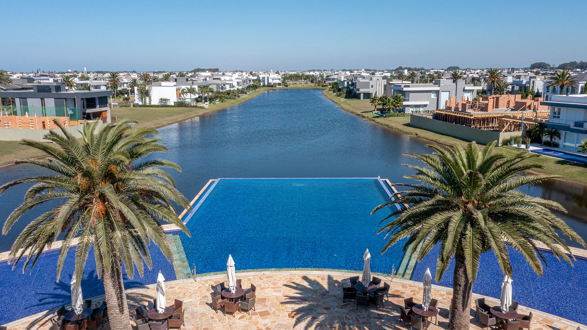 Foto do deck da piscina do club house do Enseada entre palmeiras, com o lago e casas do condomínio ao fundo