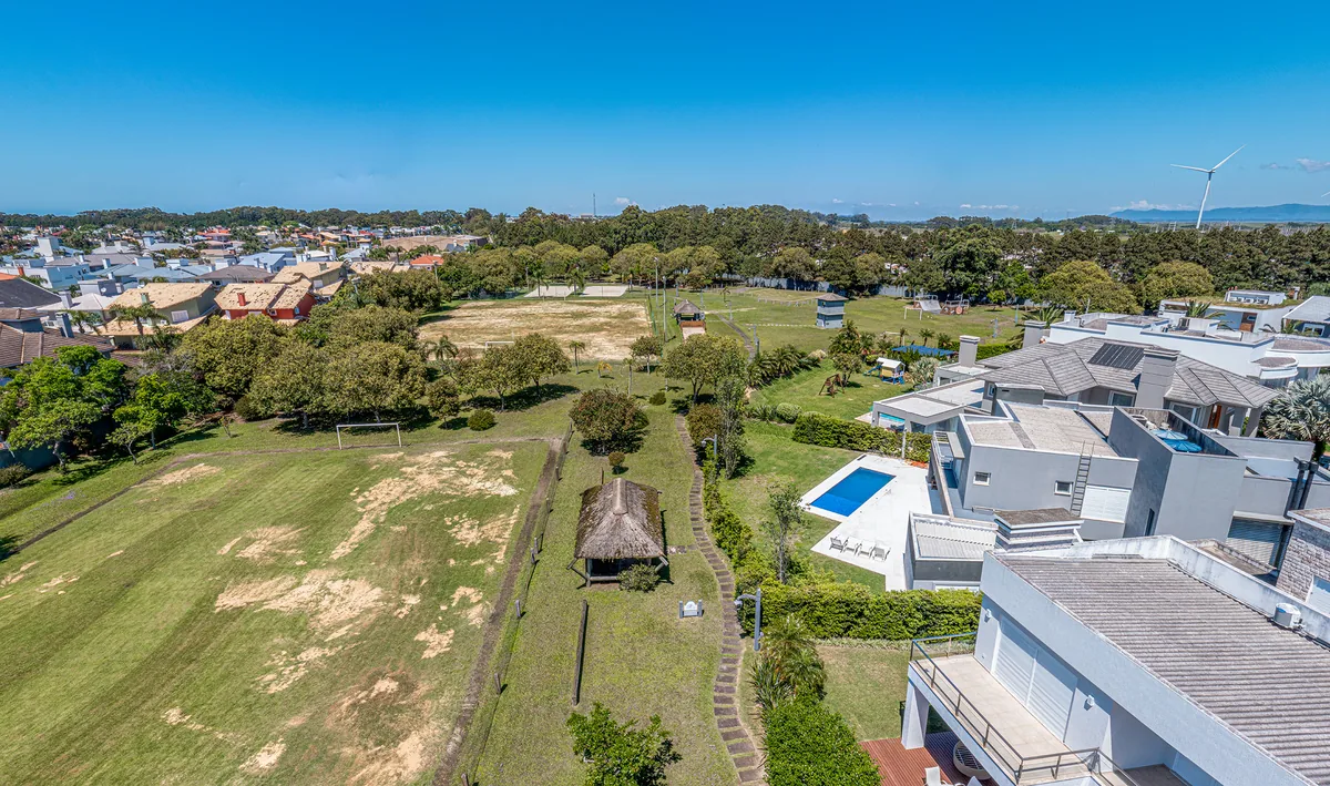 Vista aérea do campo de futebol gramado, quadras e playground do Atlântida Ilhas Park, com quiosque de telhado de sapé e aerogerador no horizonte
