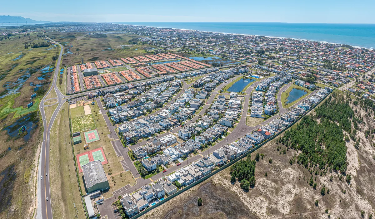 Vista aérea de uma rua interna larga e reta do Las Palmas, ladeada por casas modernas densamente construídas, com um dos lagos internos ao fundo
