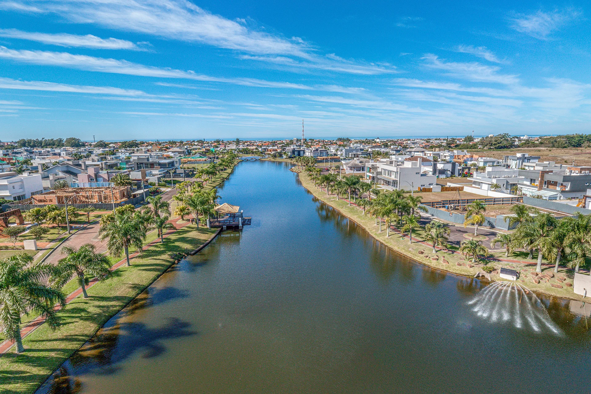 Foto aérea da ponte branca em arco sobre o canal interno do Quintas do Lago, com fonte decorativa e casas nas margens, em Xangri-Lá