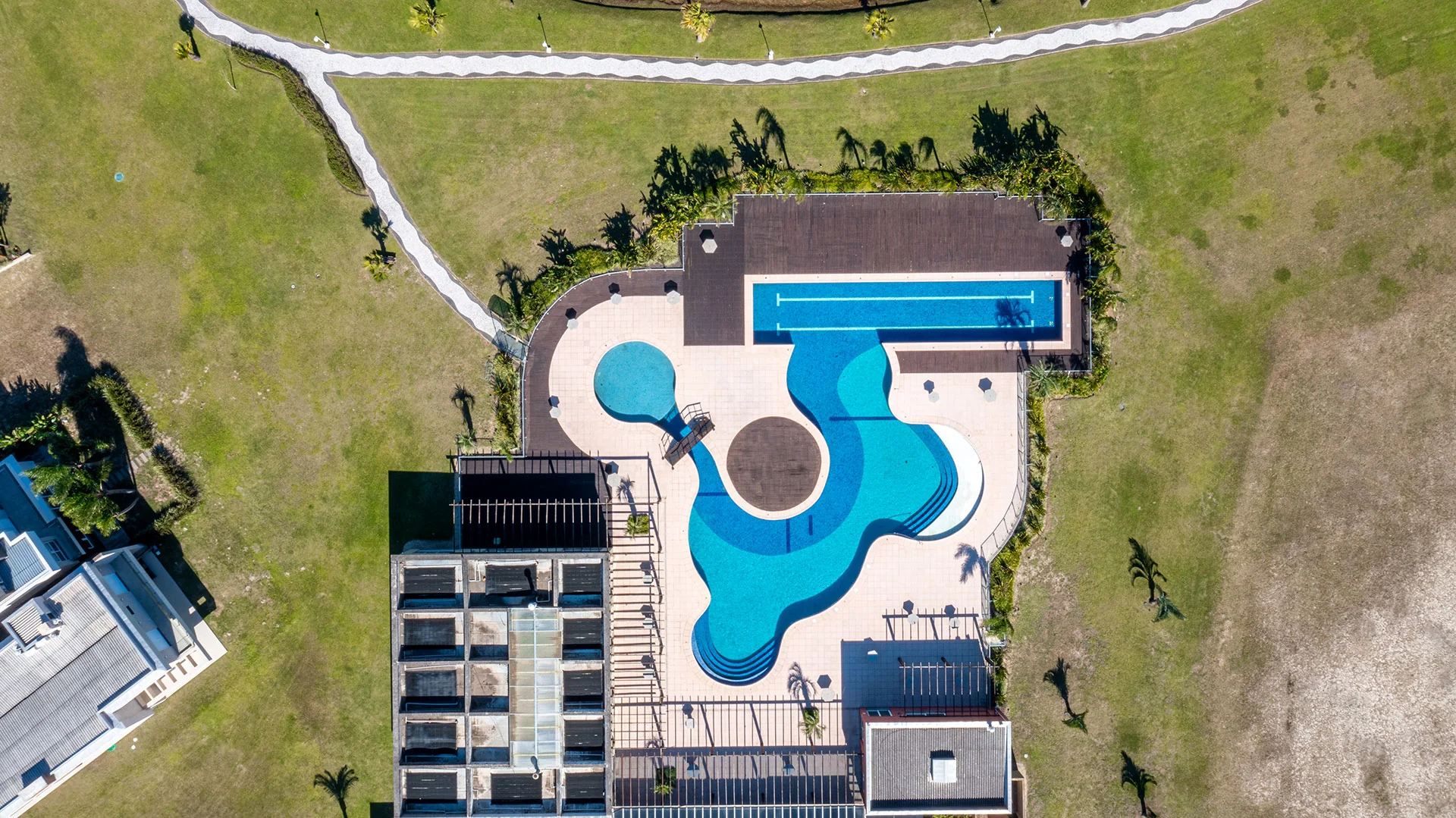 Foto aérea de cima da piscina de formato orgânico do clube do Riviera, com deck, pergolado de madeira e palmeiras