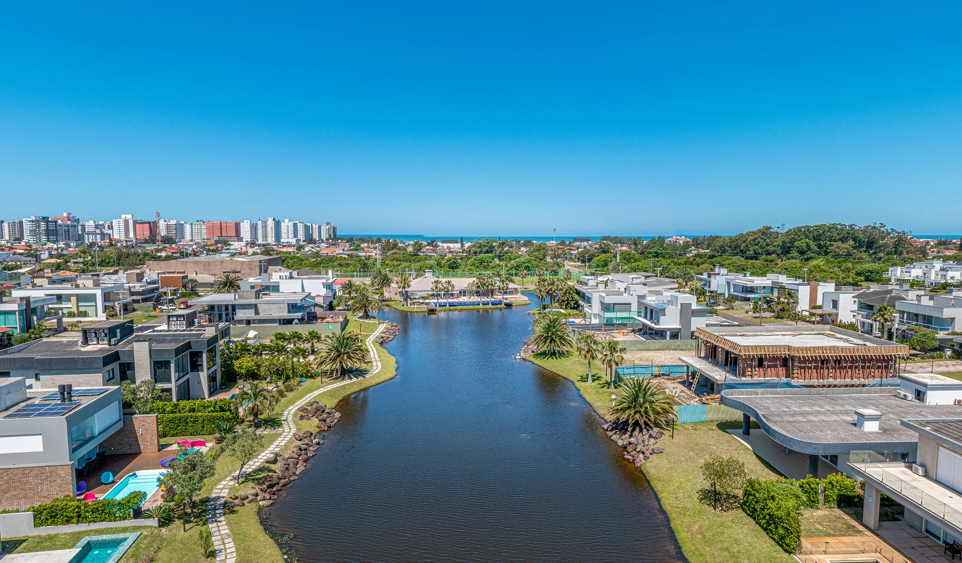 Foto aérea do canal interno do Villaggio Atlântida, com casas nas margens, caminho de pedras e skyline de Xangri-Lá ao fundo