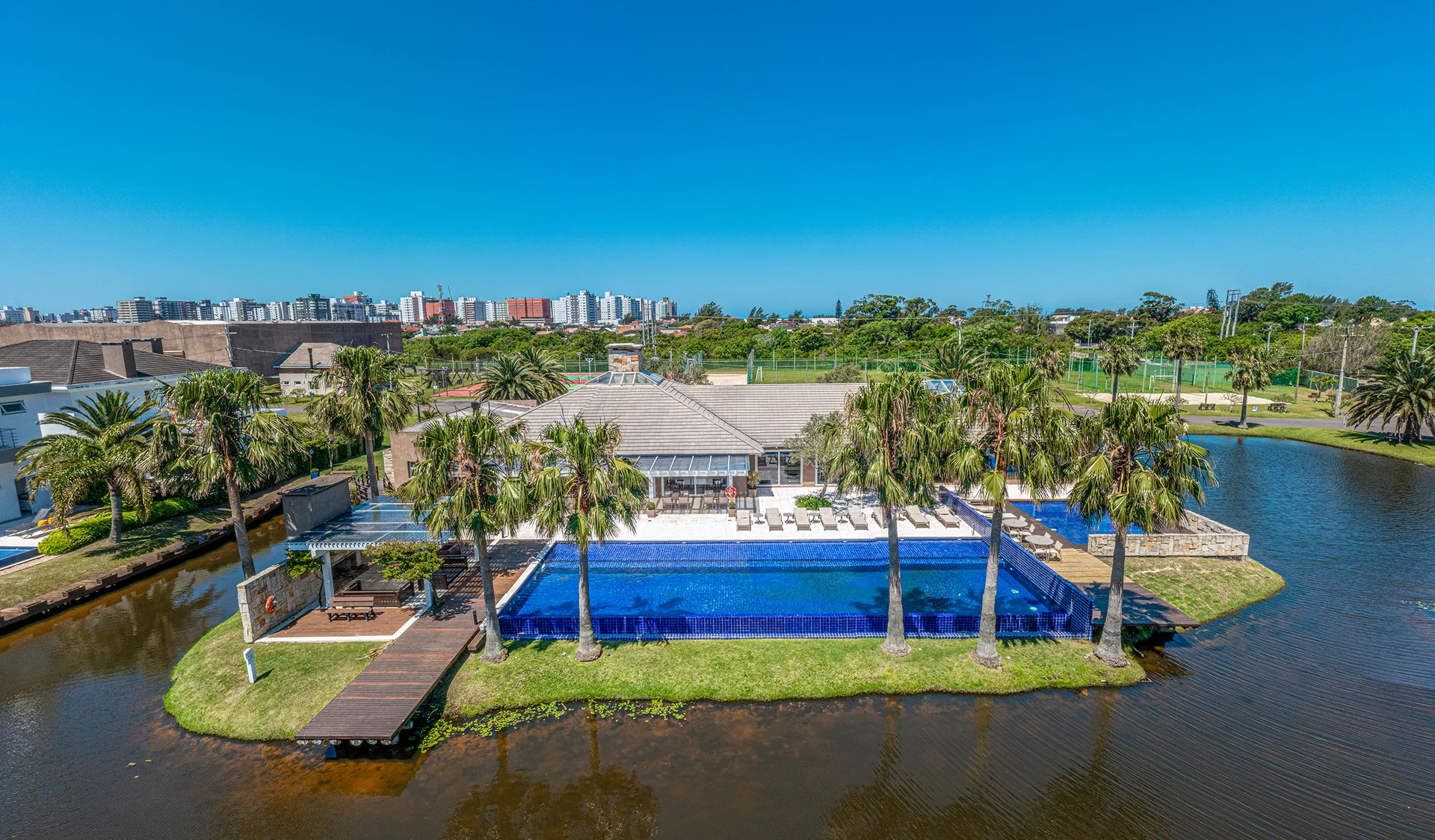 Foto aérea da piscina de borda azul do clube do Villaggio Atlântida, com deck, quiosque coberto e palmeiras sobre a ilha