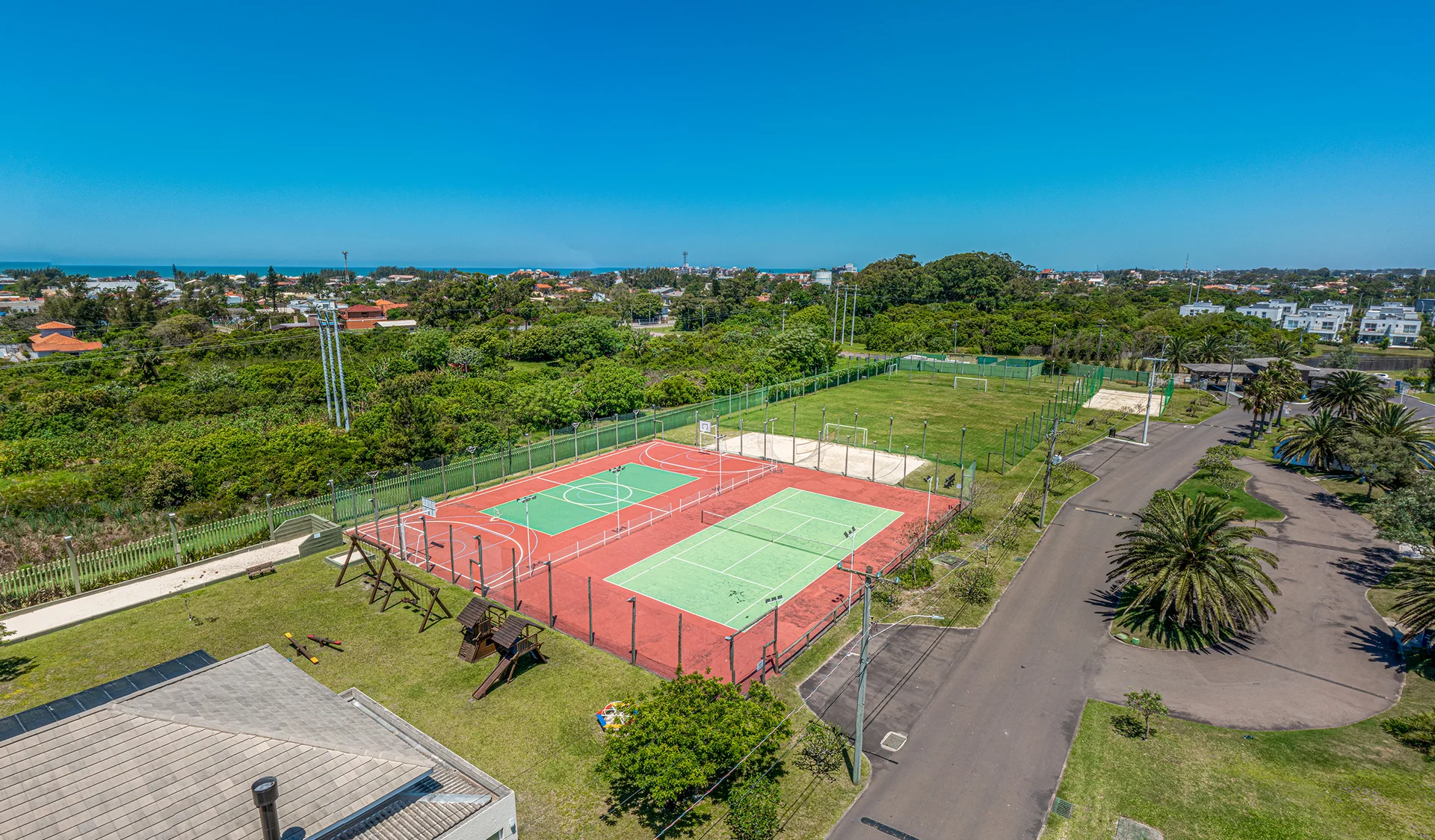 Foto aérea do conjunto esportivo do Villaggio Atlântida com campo de futebol, quadras e playground, skyline de Xangri-Lá ao fundo