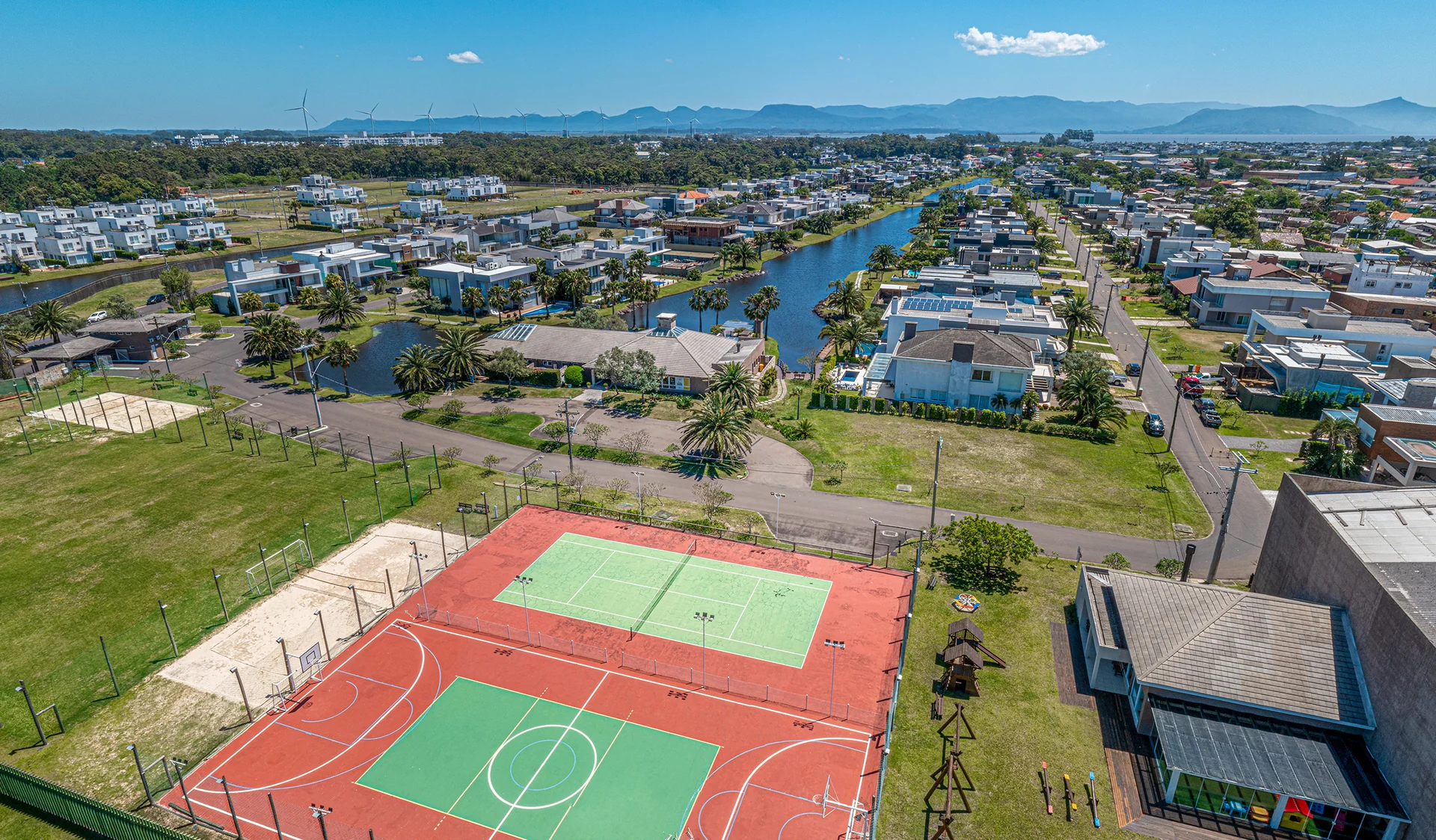 Foto aérea da quadra de tênis e da quadra poliesportiva do Villaggio Atlântida, com playground e serra ao fundo
